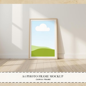 Könnte beinhalten: Ein A4-Fotorahmen-Mockup, das an einer weißen Wand lehnt. Das Kunstwerk im Inneren zeigt einen blauen Himmel, eine weiße Wolke und grüne Hügel. Der Rahmen ist aus hellem Holz. Der Text unten lautet "A4 PHOTO FRAME MOCKUP CANVA FRAME".