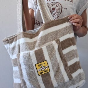 Peut inclure: Sac de plage à rayures marron et blanches, avec une poche avant et de longues anses. Le sac porte un écusson avec l'inscription "Have a Nice Day !" et des motifs floraux. Le sac est en matière douce, semblable à une serviette.