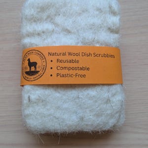 May include: A rectangular, off-white wool dish scrubbie with an orange band. The band reads "Natural Wool Dish Scrubbies," highlighting features like "Reusable," "Compostable," and "Plastic-Free." The scrubbie is made of natural fiber.