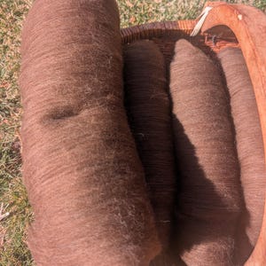 May include: A brown, cylindrical bundle of wool sits in a woven basket with a wooden handle. Several smaller, similar bundles are also in the basket. The wool has a soft, textured appearance, and the basket is set on a grassy surface.