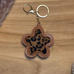 Può includere: Un portachiavi a forma di fiore con un centro con stampa leopardata e bordi marroni simili alla pelle. Il portachiavi ha un moschettone e un anello dorati. Lo sfondo è una superficie di legno marrone chiaro.