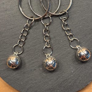 Könnte beinhalten: Drei silberne Schlüsselanhänger mit Fußball-Anhängern. Jeder Schlüsselanhänger hat einen großen Ring, eine Kette und einen kleinen Fußball-Anhänger. Die Fußbälle haben ein detailliertes, strukturiertes Design. Die Schlüsselanhänger liegen auf einer dunkelgrauen Oberfläche.