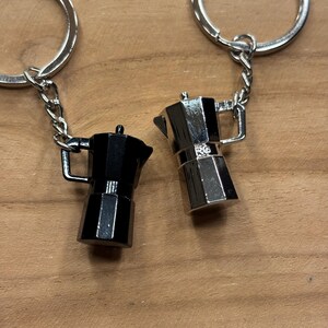 Può includere: Due portachiavi a forma di caffettiera in miniatura. Uno è nero, l'altro argentato. Entrambi sono attaccati a una catena d'argento e a un portachiavi d'argento. I portachiavi sono su una superficie di legno.