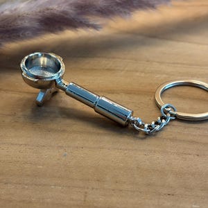 Può includere: Un portachiavi a forma di portafiltro per macchina da caffè espresso color argento. Il portachiavi presenta una replica in miniatura dettagliata di un portafiltro, completo di manico, cestello filtro e una catena attaccata a un portachiavi. Lo sfondo è una superficie di legno.