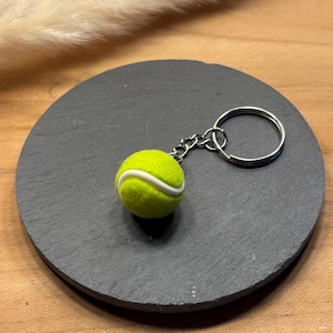 Può includere: Un portachiavi a forma di pallina da tennis. Il portachiavi presenta una piccola pallina da tennis giallo brillante con una striscia bianca, attaccata a una catena e un anello argentati. Il portachiavi è appoggiato su un sottobicchiere rotondo grigio scuro.