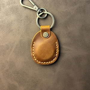 May include: A brown leather keychain with a metal clasp and ring. The leather tag is oval-shaped and stitched with a contrasting thread. The metal hardware has a silver finish. The keychain is shown on a brown textured surface.