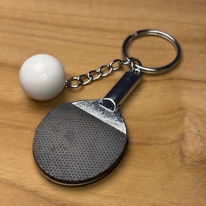 Könnte beinhalten: Ein silberfarbener Tischtennisschläger-Schlüsselanhänger mit einer schwarzen, strukturierten Oberfläche und einem weißen Ball, der mit einer Kette befestigt ist. Der Schlüsselanhänger hat einen silbernen Ring.