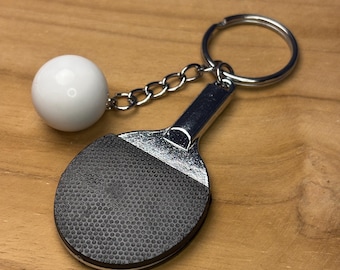 May include: A silver-colored ping pong paddle keychain with a black textured surface and a white ball attached by a chain. The keychain has a silver ring for attaching to keys or a bag.