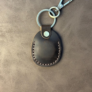 Könnte beinhalten: Ein dunkelbrauner Leder-Schlüsselanhänger mit einem silberfarbenen Karabiner und Schlüsselring. Das Lederstück ist oval geformt und an den Rändern genäht. Eine silberfarbene Niete befestigt das Leder am Schlüsselring.