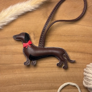 Könnte beinhalten: Ein brauner Dackel-Schlüsselanhänger aus Leder mit roter Schleife und schwarzen Akzenten. Der Schlüsselanhänger hat einen langen, geschlungenen Riemen und liegt auf einem Holzgrund. Der Hund ist etwa 10 cm lang.