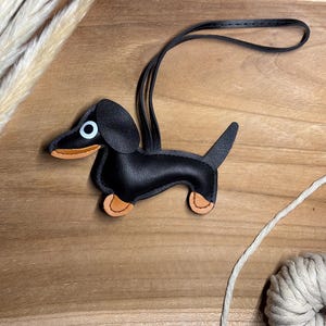 Könnte beinhalten: Ein schwarzer Dackel-Schlüsselanhänger aus Leder mit hellbraunen Pfoten und Schnauze. Der Schlüsselanhänger hat einen schwarzen Riemen zum Aufhängen. Der Hund liegt auf einer Holzoberfläche mit neutralem Hintergrund.