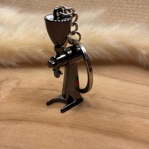 Può includere: Un portachiavi con macinacaffè nero in miniatura. Il macinacaffè ha una tramoggia, una maniglia e una base. Il portachiavi è in metallo e ha una catena e un anello argentati. Il macinacaffè è una replica dettagliata di un macinacaffè di dimensioni normali.
