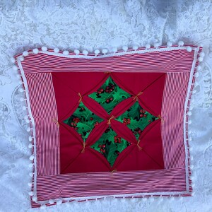 Peut inclure: Un coussin rayé rouge et blanc avec un motif de feuilles de houx au centre. Le coussin est bordé d'une garniture de pompons blancs.