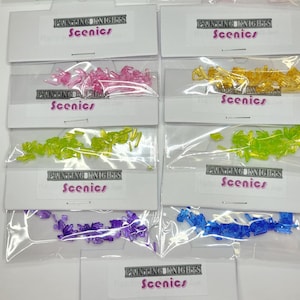 May include: A selection of miniature scenic elements in various colours, including green, red, pink, yellow, and blue. Each pack is labelled "PAINTING KNIGHTS Scenics". These small plastic pieces are likely for model building or crafting.