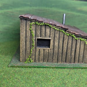 Może przedstawiać: Miniaturowa drewniana szopa z brązowym dachem i pionowym sidingiem. Widoczne jest małe, ciemne okno, a po boku rosną zielone pnącza. Szopa stoi na zielonej podstawie, z tłem zielonego pola i drzew.