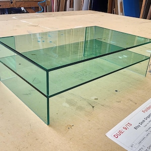 May include: A two-tiered glass coffee table with a clear green tint. The table is made of thick glass and has a modern design.