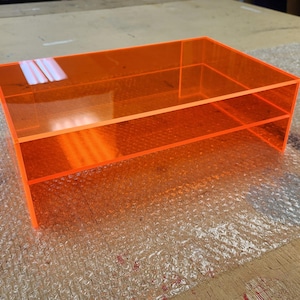 May include: A two-tiered orange acrylic shelf unit. The shelf is made of clear acrylic with a bright orange tint.