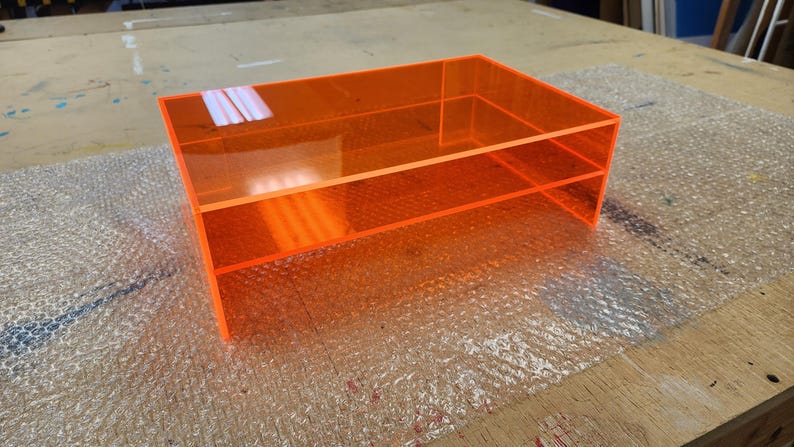 May include: A two-tiered orange acrylic shelf unit. The shelf is made of clear acrylic with a bright orange tint.
