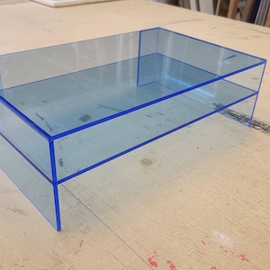 May include: A two-tiered acrylic shelf unit with a blue tint and blue edging. The shelf unit is made of clear acrylic and has a modern, minimalist design.