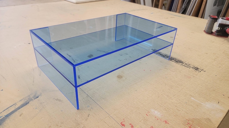 May include: A two-tiered acrylic shelf unit with a blue tint and blue edging. The shelf unit is made of clear acrylic and has a modern, minimalist design.
