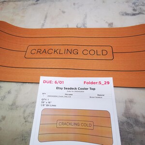 May include: A brown seadeck cooler top with the text "CRACKLING COLD" printed on it. The cooler top is 39 inches by 16 inches and has 1/8 inch bit lines.