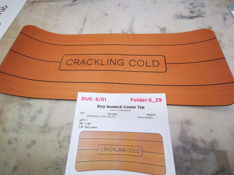 May include: A brown seadeck cooler top with the text "CRACKLING COLD" printed on it. The cooler top is 39 inches by 16 inches and has 1/8 inch bit lines.