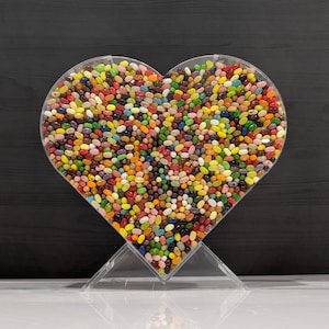 May include: A clear, heart-shaped container filled with colorful jelly beans. The container is supported by a clear stand, and the candy is a mix of various colors, including red, yellow, green, and purple.