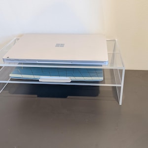 May include: A clear acrylic laptop stand with two tiers. A silver laptop is on the top tier and a blue keyboard is on the bottom tier.