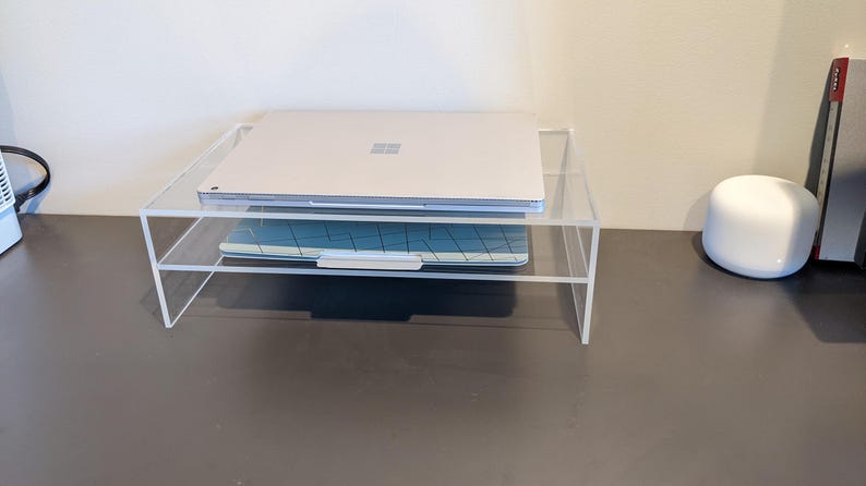 May include: A clear acrylic laptop stand with two tiers. A silver laptop is on the top tier and a blue keyboard is on the bottom tier.