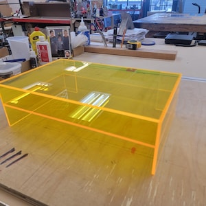 May include: A bright yellow acrylic box with a clear top and a shelf inside. The box is sitting on a wooden surface.