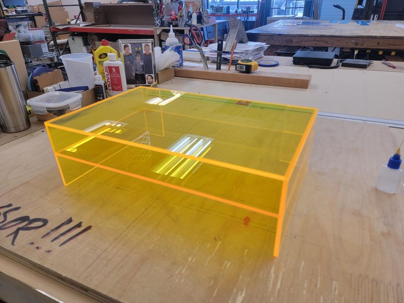 May include: A bright yellow acrylic box with a clear top and a shelf inside. The box is sitting on a wooden surface.
