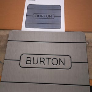 May include: A gray, rectangular, non-slip surface with the word "BURTON" printed in black. The surface is 14.43 inches by 11.71 inches and has 1/8 inch bit lines.