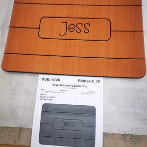 May include: A brown wooden cooler top with the name "Jess" engraved in the center. The top is 19.125 inches by 13.675 inches and has 1/8 inch bit lines.