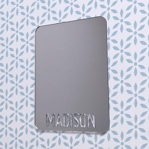 Puede incluir: Un espejo plateado con el nombre "MADISON" grabado en la parte inferior.