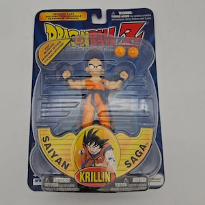 May include: A Dragon Ball Z Krillin action figure in original packaging. The figure wears an orange gi with a blue belt and wristbands. Included are two small orange spheres with red stars. The card features the text Saiyan Saga and Krillin.