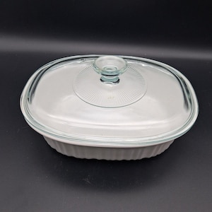 May include: An oval white ceramic casserole dish with vertical fluted sides and a clear glass lid. The lid features a central circular knob handle surrounded by a radiating sunburst pattern etched into the glass surface, providing a clear view.