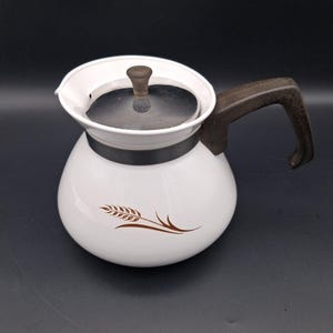 May include: A white enamel teapot featuring a brown wheat stalk graphic on the side. The teapot has a dark brown handle, a metallic silver lid with a small brown knob, and a wide rim. The design is simple with a rounded body and a spout.