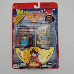 May include: A Dragon Ball Z Babidi action figure in original blister packaging. The set includes a Babidi figure with a blue skirt and red cape, a small pink cocoon egg, three orange dragon balls, and a collectible card featuring Shenron.