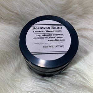 May include: A round, black container of beeswax balm with a white label. The label reads "Beeswax Balm Lavender Thyme Scent" and lists ingredients. The net weight is 1 oz.