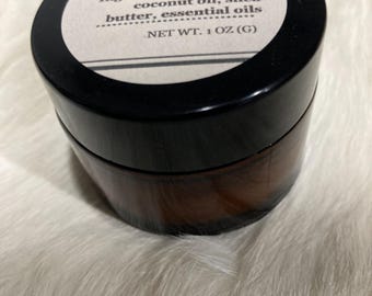 May include: A small, brown jar with a black lid and a white label that reads "Beeswax Balm". The label lists the ingredients: beeswax, coconut oil, shea butter, and essential oils. The jar contains 1 ounce of balm.