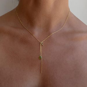 Könnte beinhalten: Eine goldene Kettenkette mit einem kleinen grünen Edelsteinanhänger und einem zarten Goldanhänger. Die Kette wird von einem Model getragen.