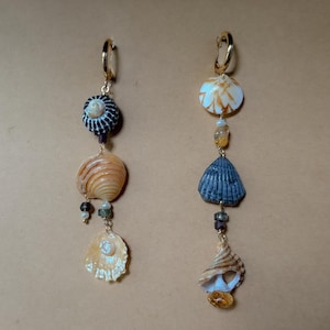 Könnte beinhalten: Ein Paar Ohrhänger mit natürlichen Muscheln und kleinen Perlen. Die Ohrringe haben goldfarbene Creolen und bestehen aus verschiedenen Muschelformen in Weiß, Beige und Dunkelblau. Die Perlen sind in Braun- und Grüntönen gehalten.