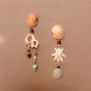 Puede incluir: Un par de pendientes colgantes con conchas naturales, pequeñas cuentas de color rojo oscuro, perlas color crema y cuentas cuadradas de color verde claro en cadenas de metal dorado. El pendiente izquierdo incluye un fragmento de concha blanca texturizada, mientras que el derecho presenta una concha en forma de estrella y una concha lisa, redonda y clara en la base.