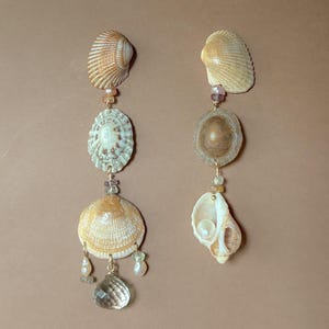 May include: A pair of long drop earrings featuring natural seashells in shades of tan, beige, and cream. The earrings are accented with small, multi-coloured gemstone beads, tiny white pearls, and a clear, faceted teardrop-shaped crystal pendant.