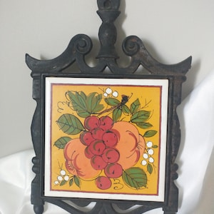 Op de afbeelding: Een zwarte gietijzeren trivet met een vierkante tegelinzet met een fruitontwerp. De tegel heeft een gele achtergrond met illustraties van rode kersen, oranje perziken en groene bladeren. De trivet heeft een decoratief handvat.