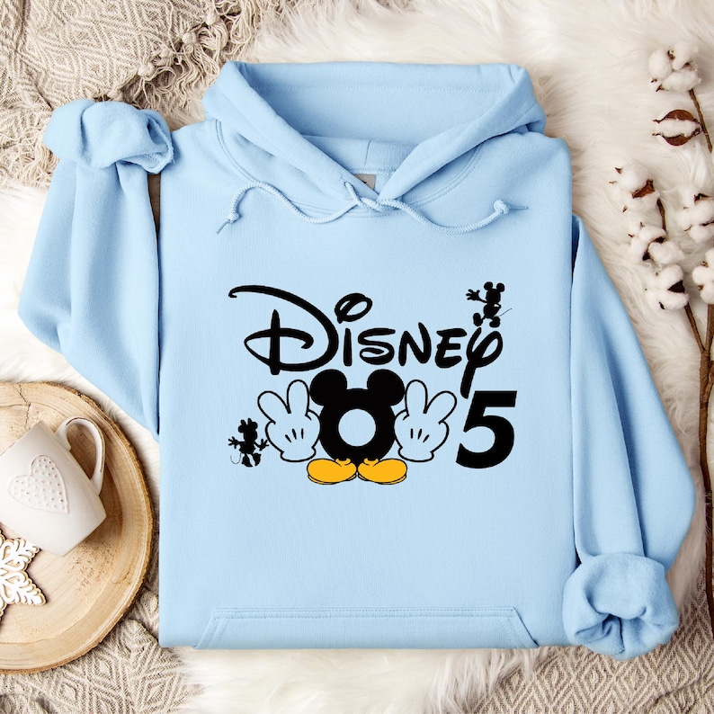 Op de afbeelding: Lichtblauwe hoodie met een zwarte afbeelding van Mickey Mouse en de tekst "Disney 5".