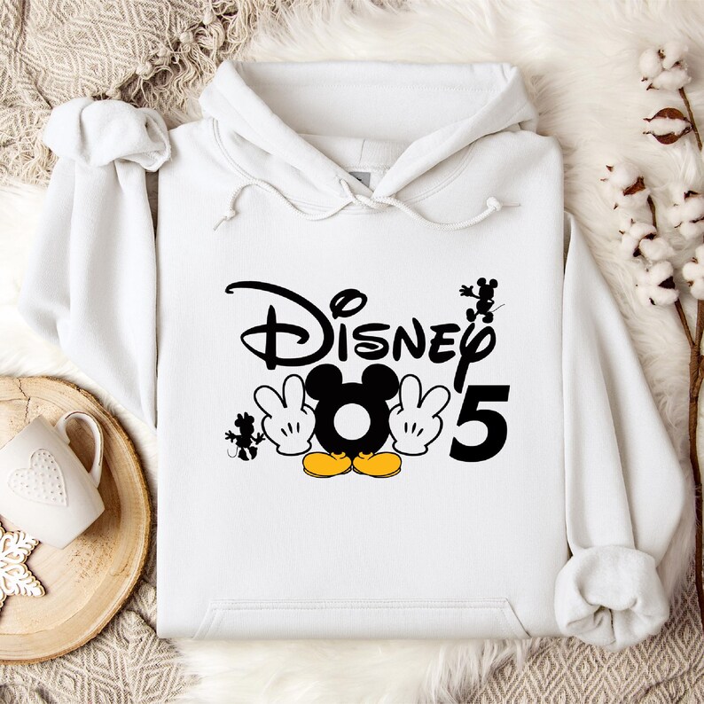 Op de afbeelding: Witte hoodie met zwarte tekst die "Disney" en een zwart nummer "5" zegt. Een zwart-witte cartoon Mickey Mouse bevindt zich in het midden van de tekst met gele schoenen.