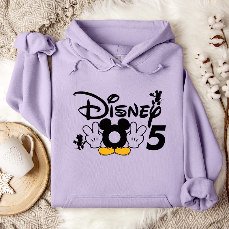 Op de afbeelding: Een lavendelkleurige hoodie met een zwart-witte afbeelding van Mickey Mouse en het woord "Disney" met het nummer "5" ernaast.
