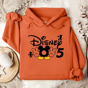 Op de afbeelding: Oranje hoodie met een zwarte afbeelding van Mickey Mouse en de tekst "Disney 5".