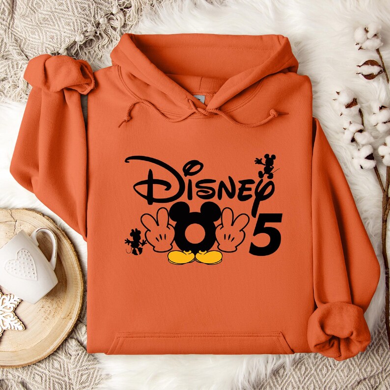 Op de afbeelding: Oranje hoodie met een zwarte afbeelding van Mickey Mouse en de tekst "Disney 5".